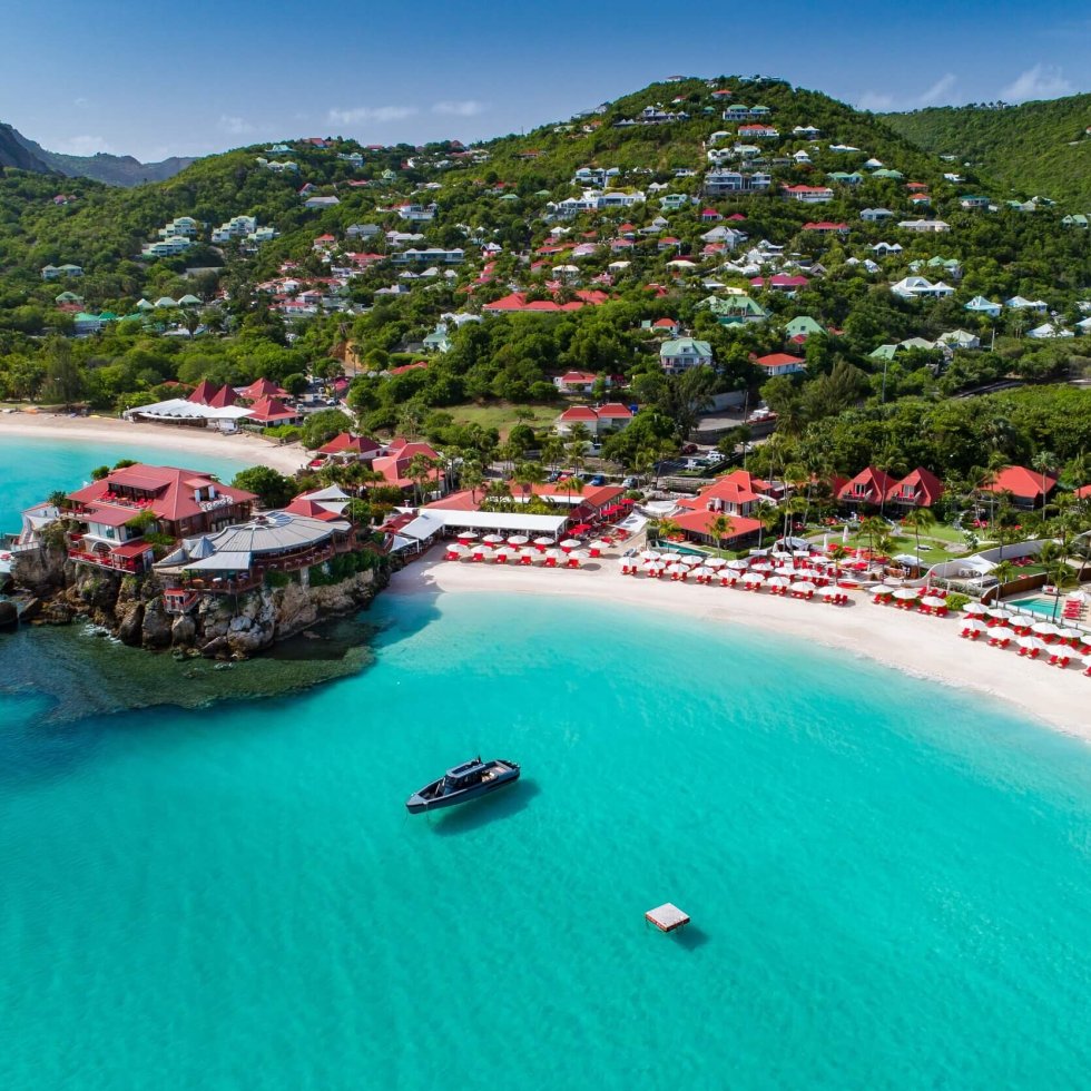 An aerial view of St. Jean Bay in St. Barths, with the iconic Eden Rock Hotel perched on its rocky outcrop, surrounded by turquoise waters and white sandy beaches