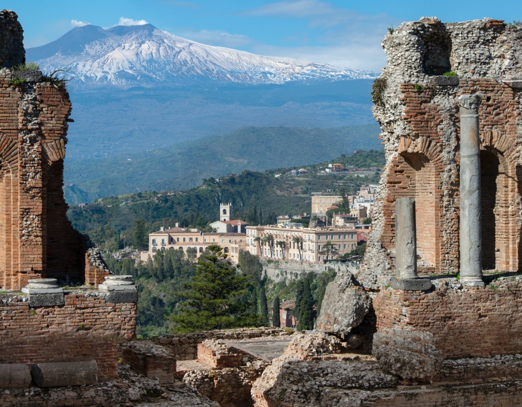 View of San Domenico Palace, Sicily’s best luxury hotel and The White Lotus filming location, seen from the ancient Greek Theatre of Taormina with Mount Etna in the background.