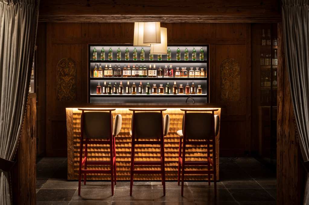 A cozy bar area with three high stools and a wooden counter, behind which a backlit shelf displays an array of various bottles.