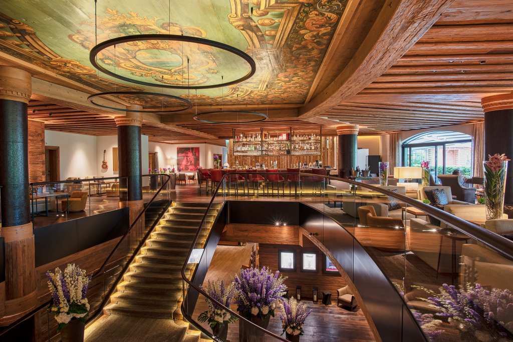 Interior view of The Alpina Gstaad hotel, showcasing a beautifully designed space with wooden accents, elegant lighting, and a vibrant ceiling mural.