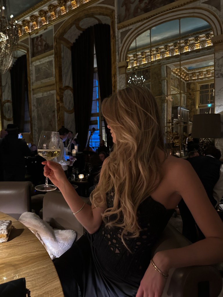 A guest enjoying a glass of champagne at the marble bar inside Bar Les Ambassadeurs at Hôtel de Crillon in Paris, with warm ambient lighting reflecting off the polished surface, elegant bar stools, and a refined, intimate luxury hotel bar atmosphere in the background.