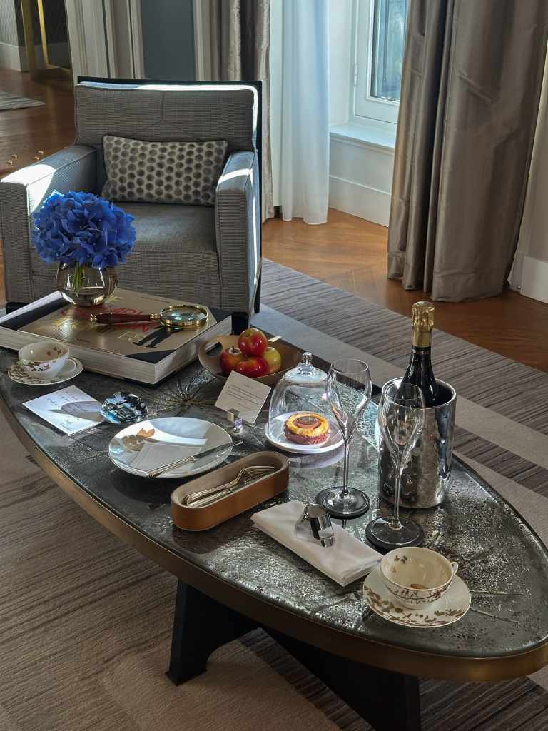 In-suite welcome setup featuring a chilled bottle of champagne, fresh floral arrangement, and assorted pastries presented on a table inside a luxury hotel room, arranged as a personalized arrival amenity.