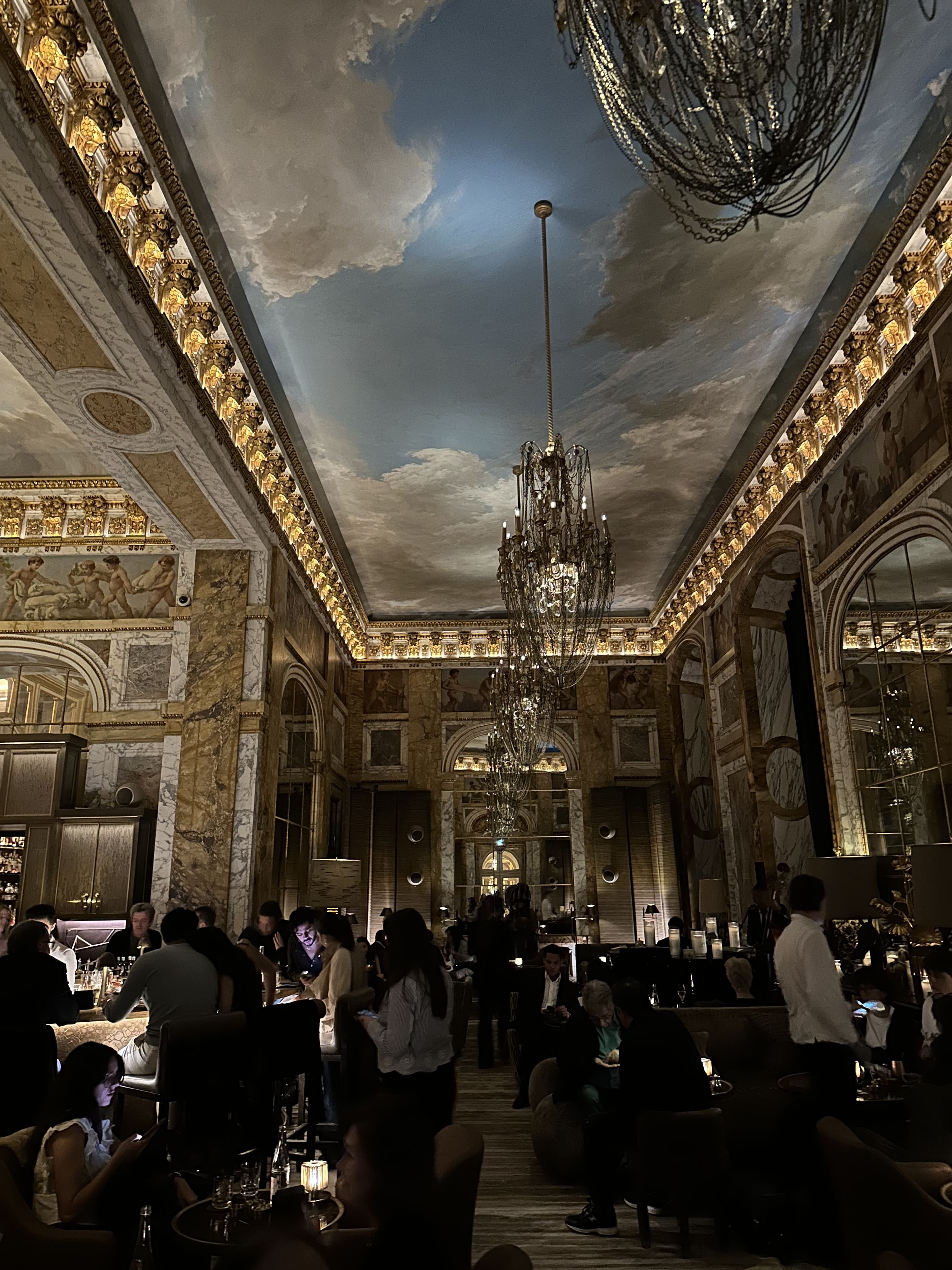 Busy evening inside Bar Les Ambassadeurs at Hôtel de Crillon in Paris, with guests seated at the illuminated marble bar and plush armchairs, soft golden lighting reflecting off mirrored walls and chandeliers, and bartenders moving through a lively, elegant crowd in a refined hotel bar atmosphere.