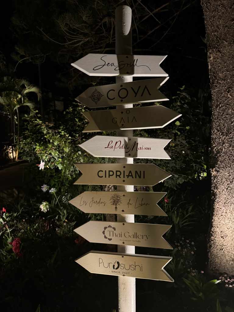 A tall signpost with multiple white arrow signs pointing in different directions, each labeled with restaurant names (e.g., Coya, Cipriani, La Petite Maison) at night, surrounded by greenery.