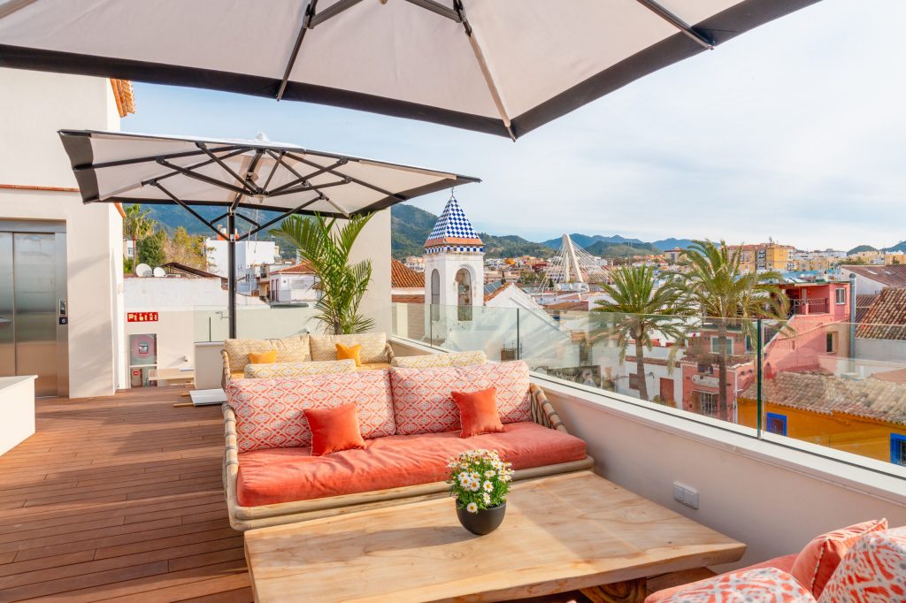 Rooftop terrace at La Fonda Heritage Hotel in Marbella showing a coral sofa with orange cushions, a wooden table, large umbrellas, and views of Marbella's old town with palm trees and colorful rooftops.